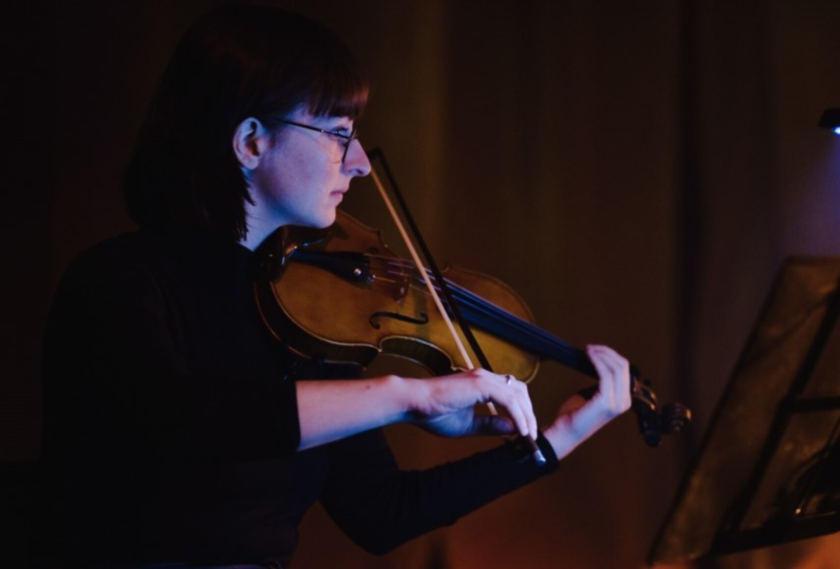 Photo en clair-obscur de la violoniste du spectacle "Hercule et Naïade" jouant du violon. Elle est de profil tournée vers la droite et son visage et ses mains sont éclairés d'une lumière bleu-mauve.