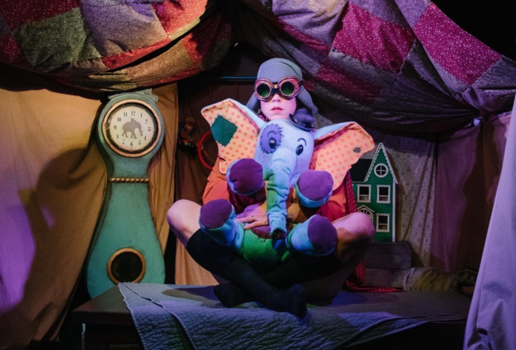 Un personnage avec des lunettes d'aviateur tient dans ses bras son toutou éléphant. Il est assis dans un décor coloré rappelant une "cabane de couvertures".