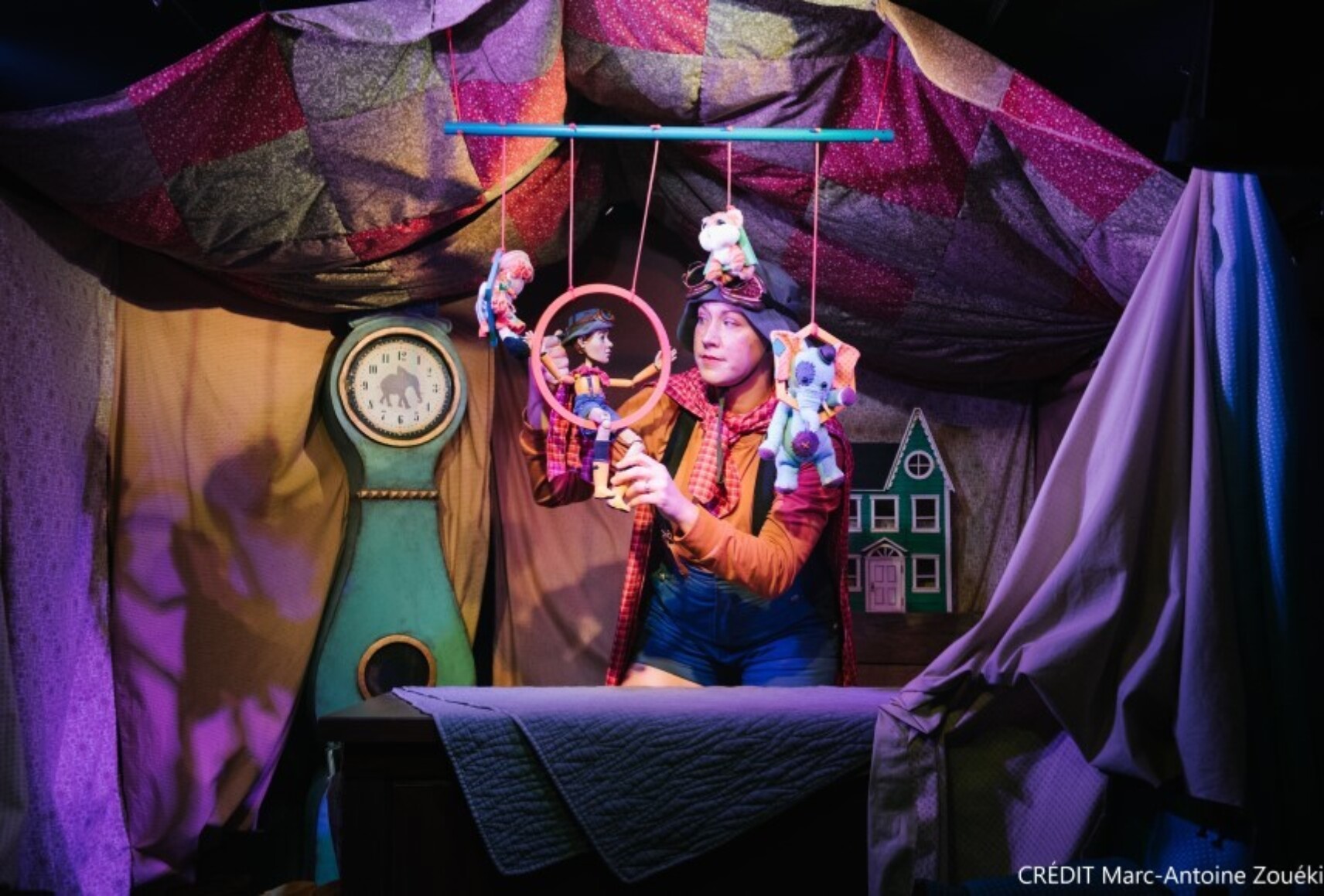 Un interprète manipule des marionnettes dans un décor coloré rappelant une "cabane de couvertures". En arrière plan, une horloge et une petite maison.
