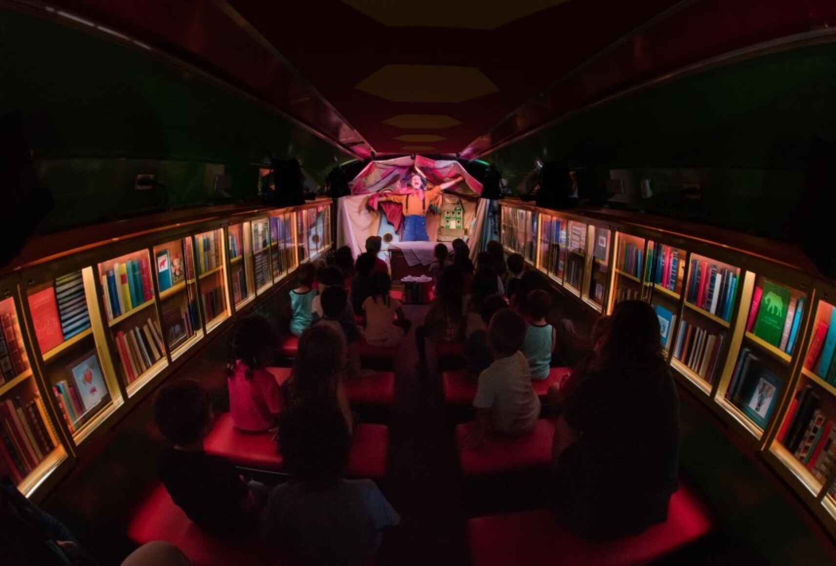 Vue d'ensemble de l'intérieur de l'autobus, transformé en théâtre. De chaque côté, les murs sont recouverts de livres dans des vitrines. Un personnage est sur scène, au fond. Des enfants sont assis sur des bancs, dans l'espace central.