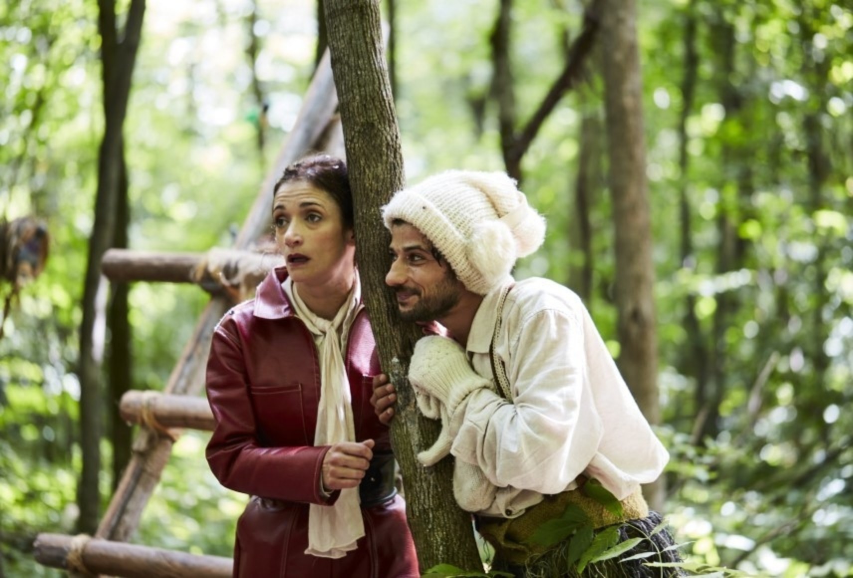 Photo en forêt de Laviat et Merlinot, l'oreille collée contre un tronc d'arbre entre eux deux. Laviat a les sourcils relevés et la bouche entrouverte, Merlinot sourit. Il porte un bonnet, une chemise et des mitaines beiges.
