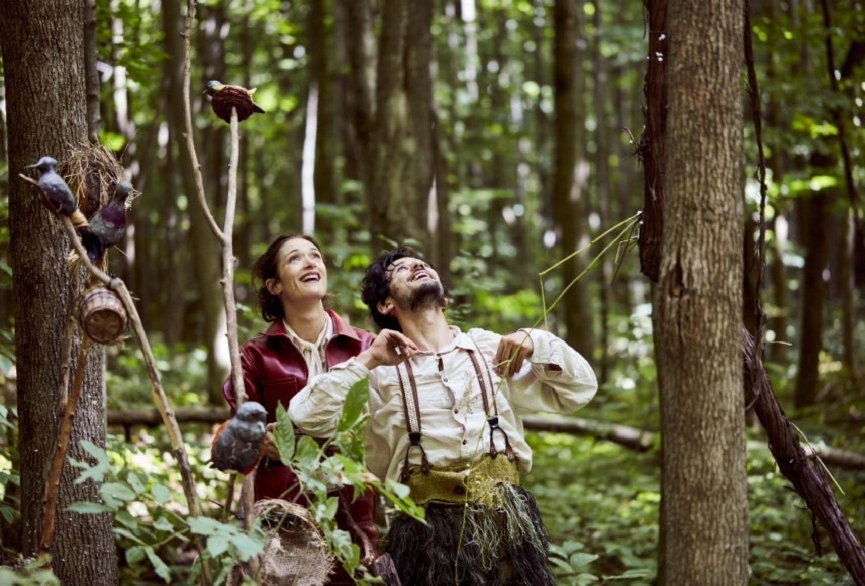 Photo dans la forêt de Laviat et Merlinot souriants, regardant vers le ciel. Des accessoires et des oiseaux en bois sont présents au premier plan.
