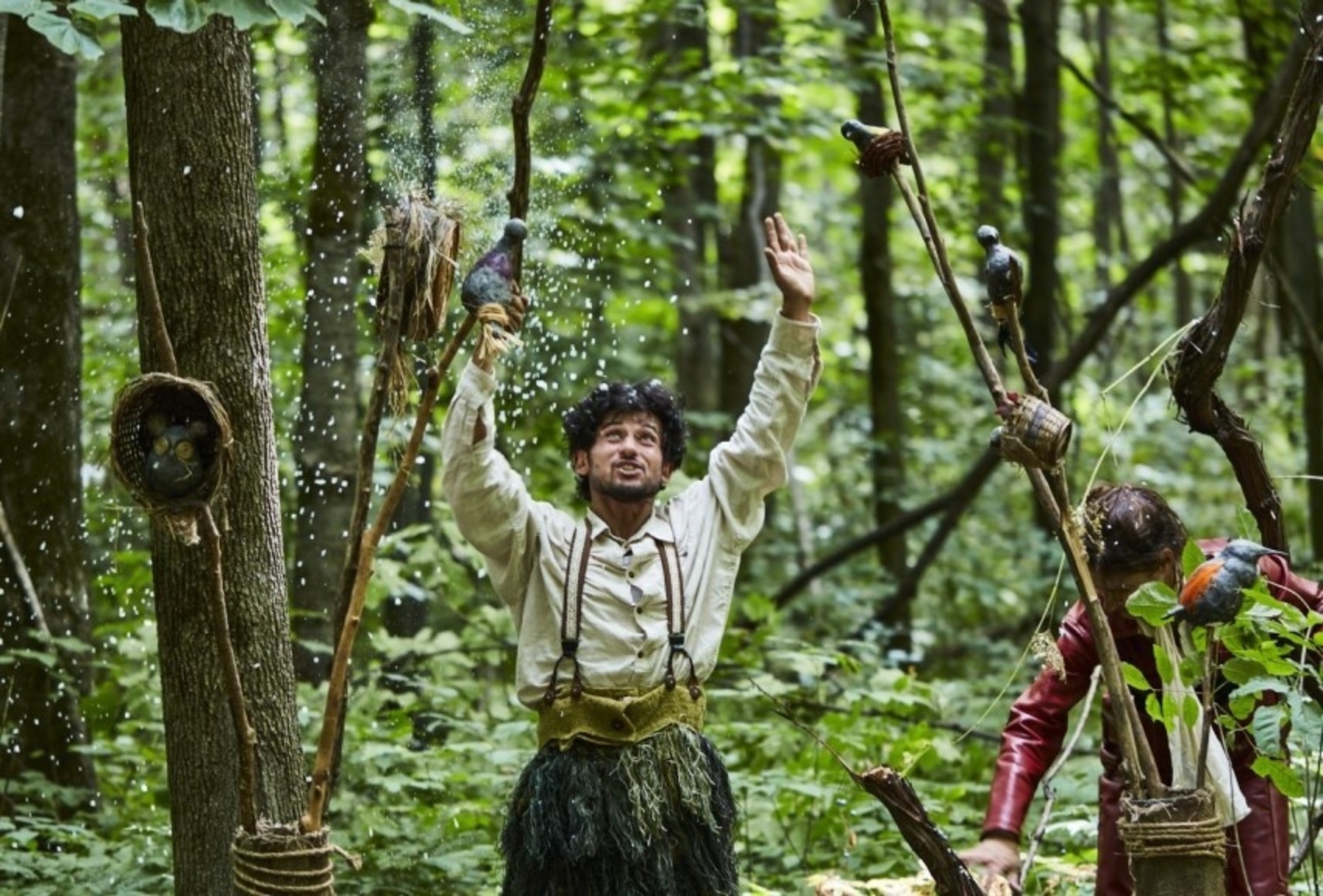 Photo dans la forêt du personnage de Merlinot jetant de la fausse neige dans les airs. Autour de lui, plusieurs accessoires et des oiseaux sculptés en bois. Il porte une chemise beige avec des bretelles et des pantalons texturés.
