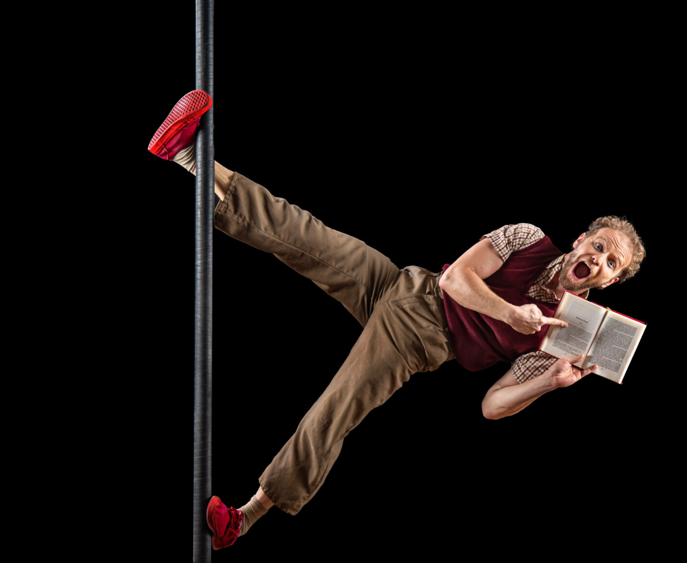 Interprête clown accroché par le bout des pieds à un mat, nous montre le contenu de son livre avec joie