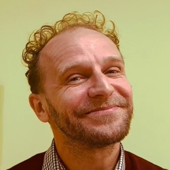 le comédien Hippolyte tout souriant. Il a la peau claire, les cheveux courts bouclés un peu roux, porte une courte barbe.