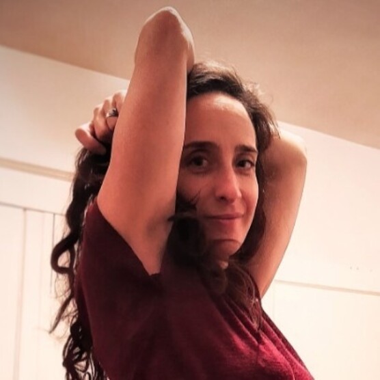 Photo portrait de l’artiste Laurence Racine, yeux bleus perçants, cheveux bruns frisés, les bras dans les airs