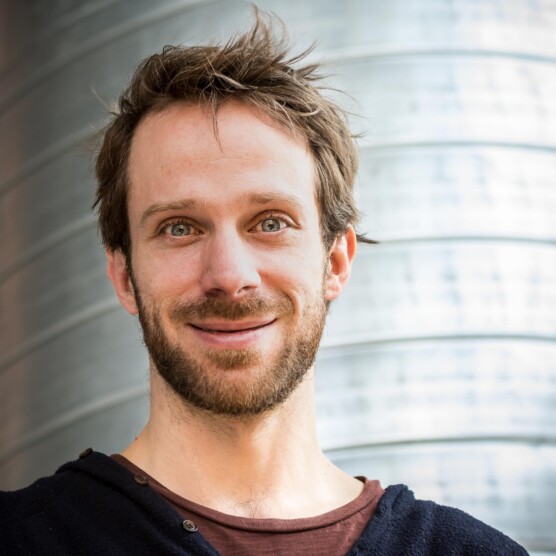 Photo portrait de l’artiste Simon Fournier, yeux bleus, cheveux châtains, regard doux, devant une colonne métallique