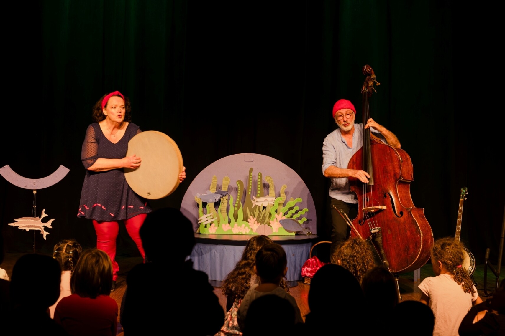 La comédienne tient un tambour de l'océan, rond et plat. Le musicien joue de la contrebasse. Entre eux, des formes découpées représentent un fond marin : des algues, des poissons, sur fond bleu.