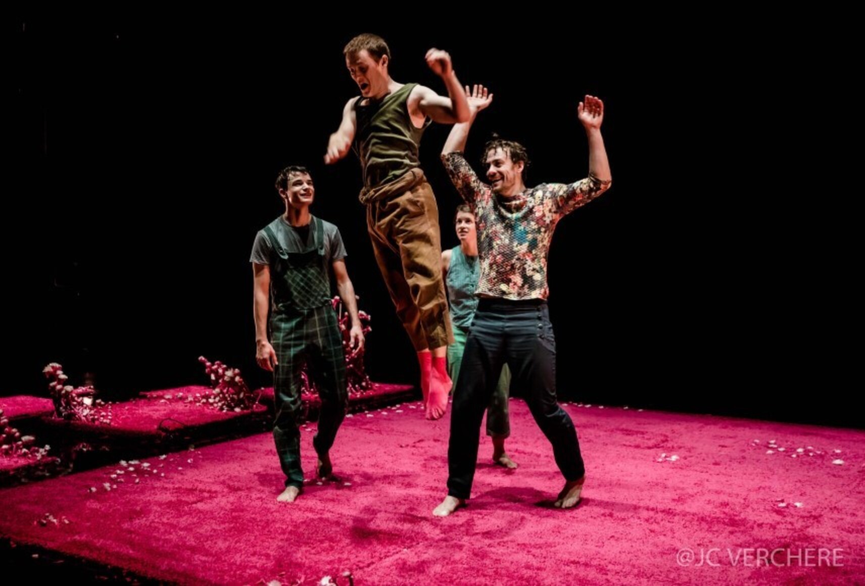 Un des personnages fait un saut au milieu des autres, également en mouvement. Ils sont sur un grand tapis rose fuchsia, devant un fond noir.
