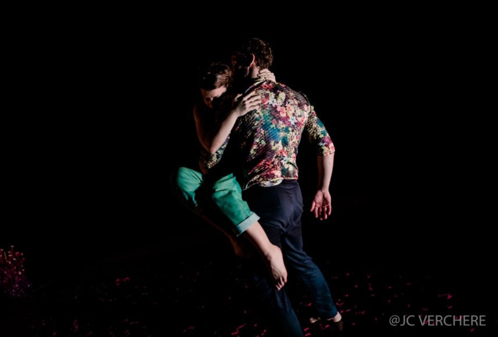 Photo en clair-obscur d'un personnage en portant un autre dans ses bras. Il est de dos et porte des pantalons marine et une chemise fleurie. On distingue les pantalons verts, une jambe et un bras de l'autre personnage.