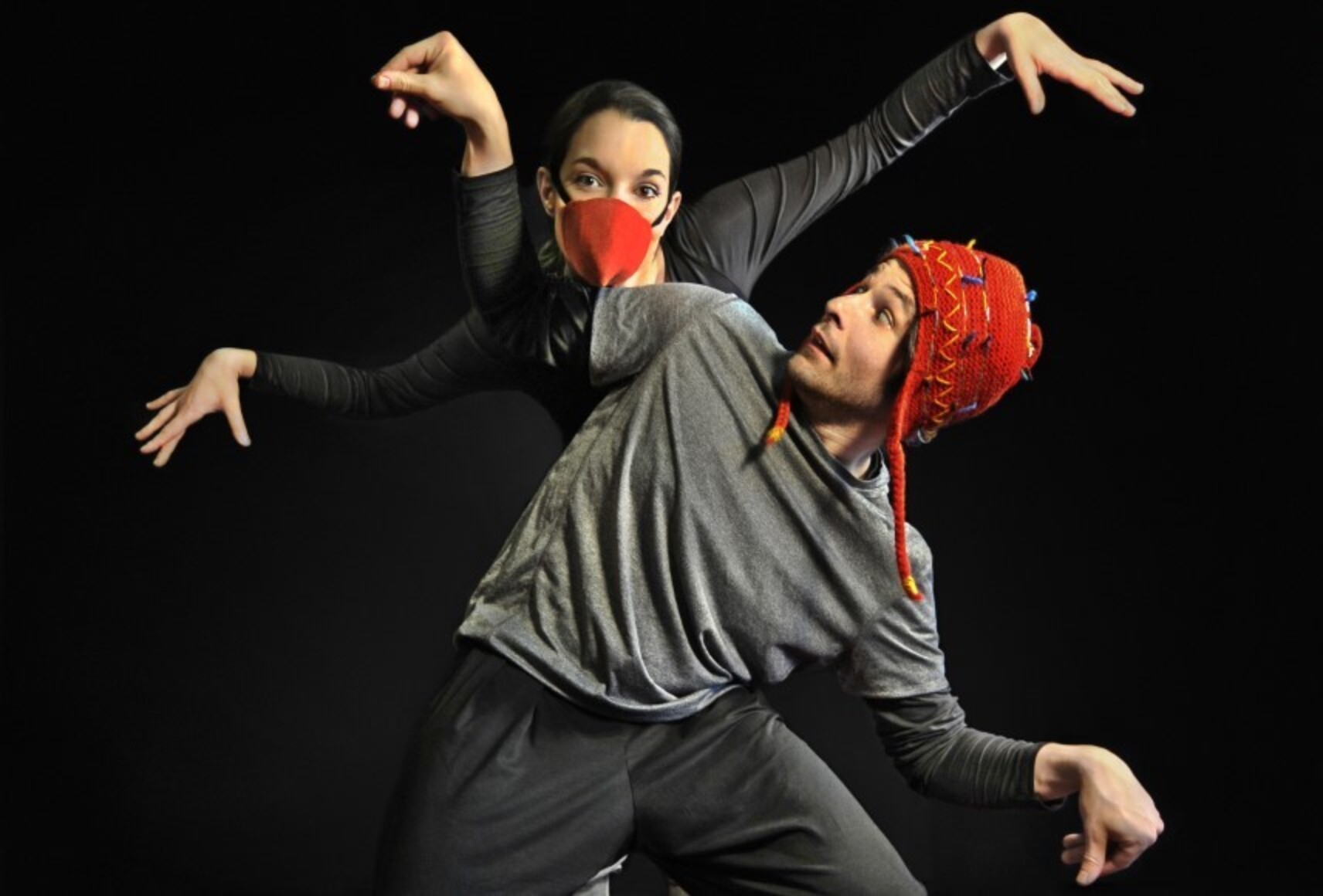 Photo des deux personnages qui bougent les bras sur un fond noir. Les deux portent des vêtements noirs et gris, l'homme une tuque rouge et regarde la femme, qui porte un masque conique rouge et regarde la caméra.