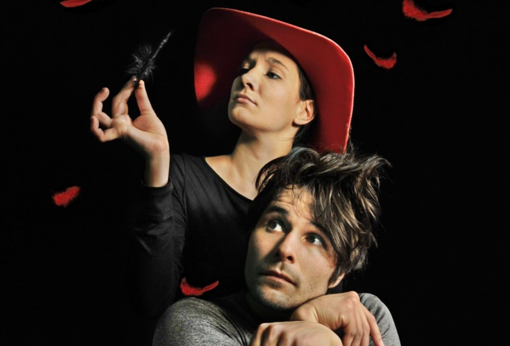 Une femme au chapeau rouge tient une plume noire dans sa main droite. Sous elle, un homme la tête appuyée sur des mains croisées, regarde vers le haut. Autour d'eux des plumes rouges tombent, sur un fond noir.