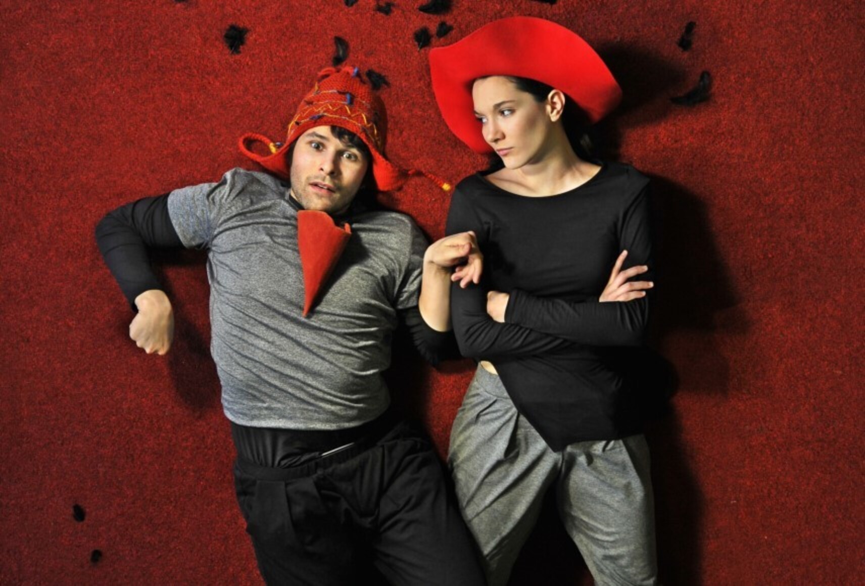 Les deux personnages sont allongés sur un sol rouge, la caméra au-dessus d'eux. À gauche un homme portant une tuque rouge, un masque conique rouge pend dans son cou. À droite, une femme au chapeau rouge le regarde, les bras croisés.