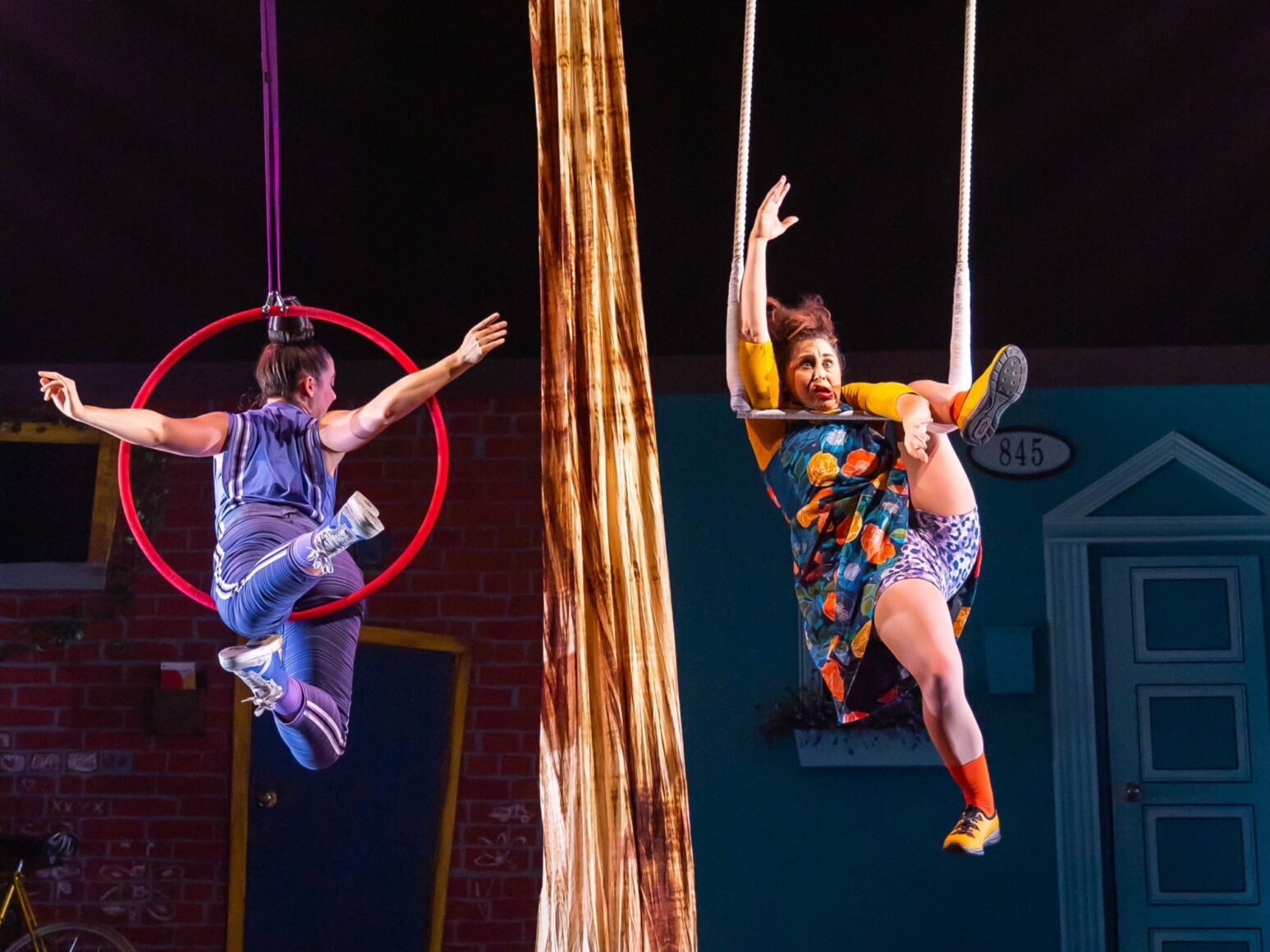 Deux artistes de cirque sont sur scène : l’une est suspendue avec grâce dans un cerceau aérien rose, l’autre semble proche de tomber, sur une balançoire fixée à un arbre stylisé au centre du décor.