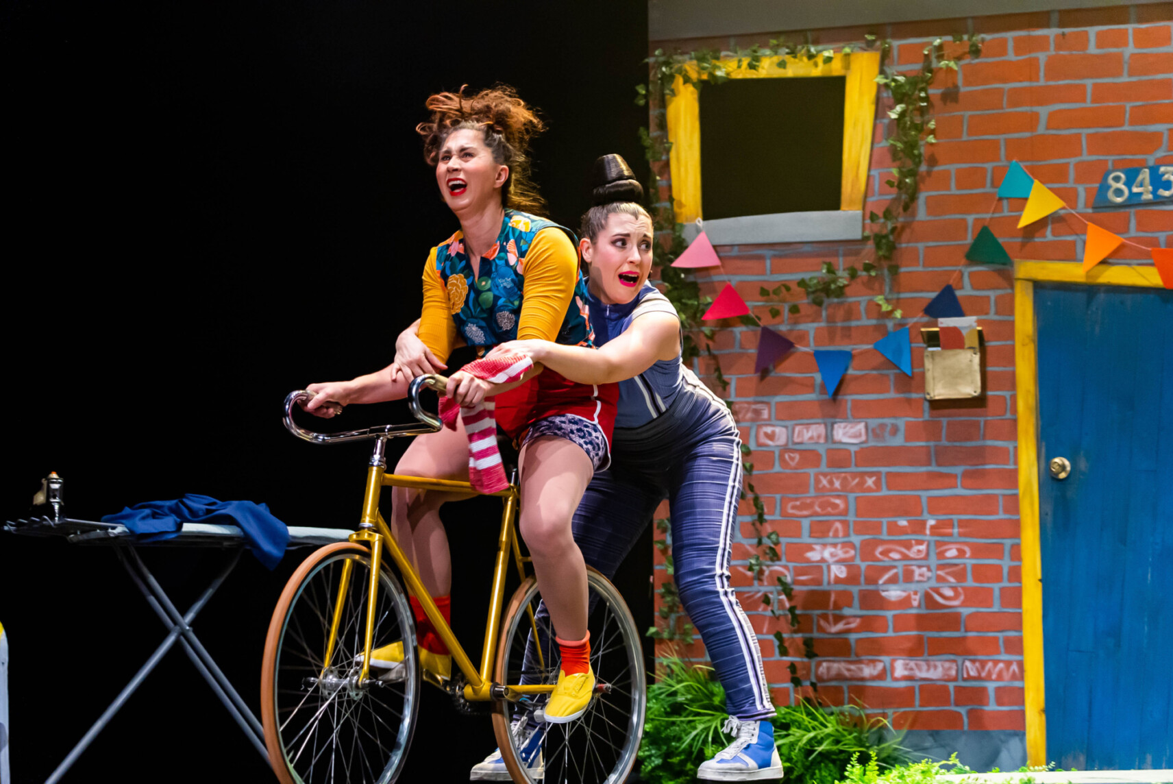 Deux artistes clownesques sur scène, l’une pédalant sur un vélo jaune, l’autre agrippée derrière elle, dans un décor coloré de façade en briques.