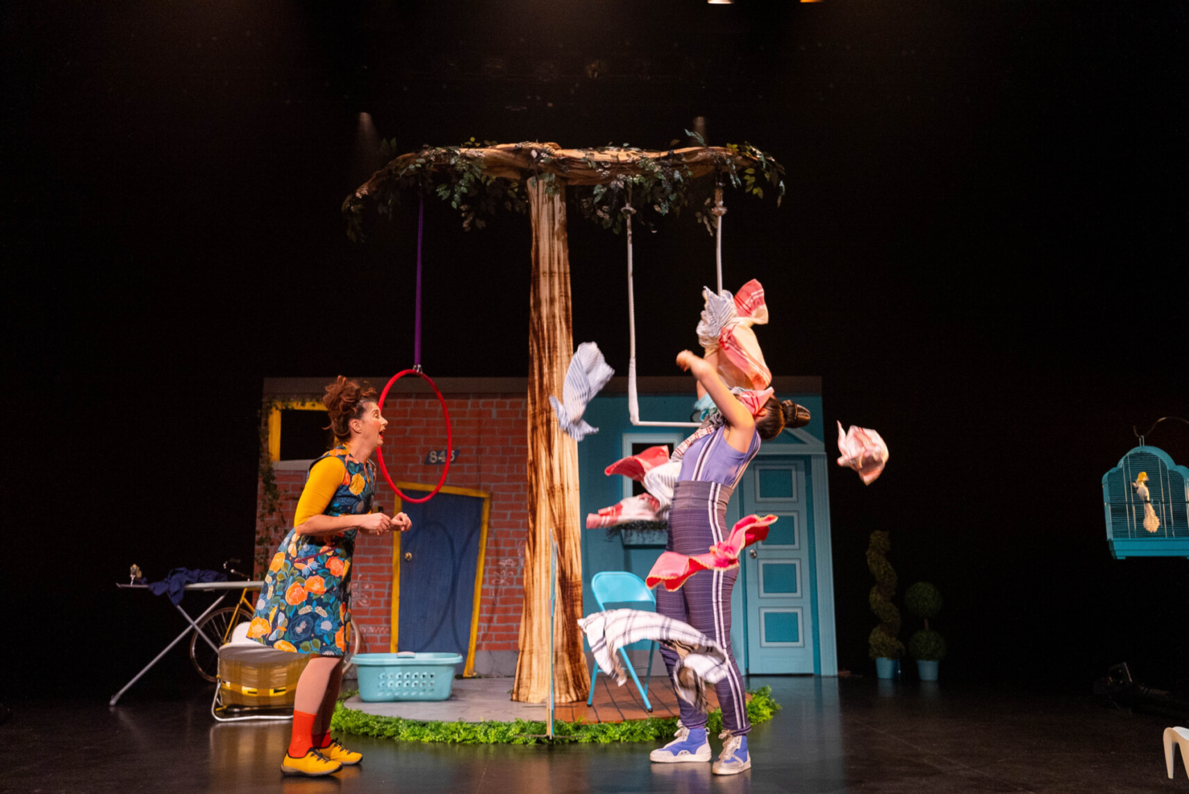 Deux comédiennes se font face, celle de gauche en robe à motifs fleurs, celle de droite regarde vers le haut et est empêtrée dans un amas de linge qui semble lui tomber dessus.