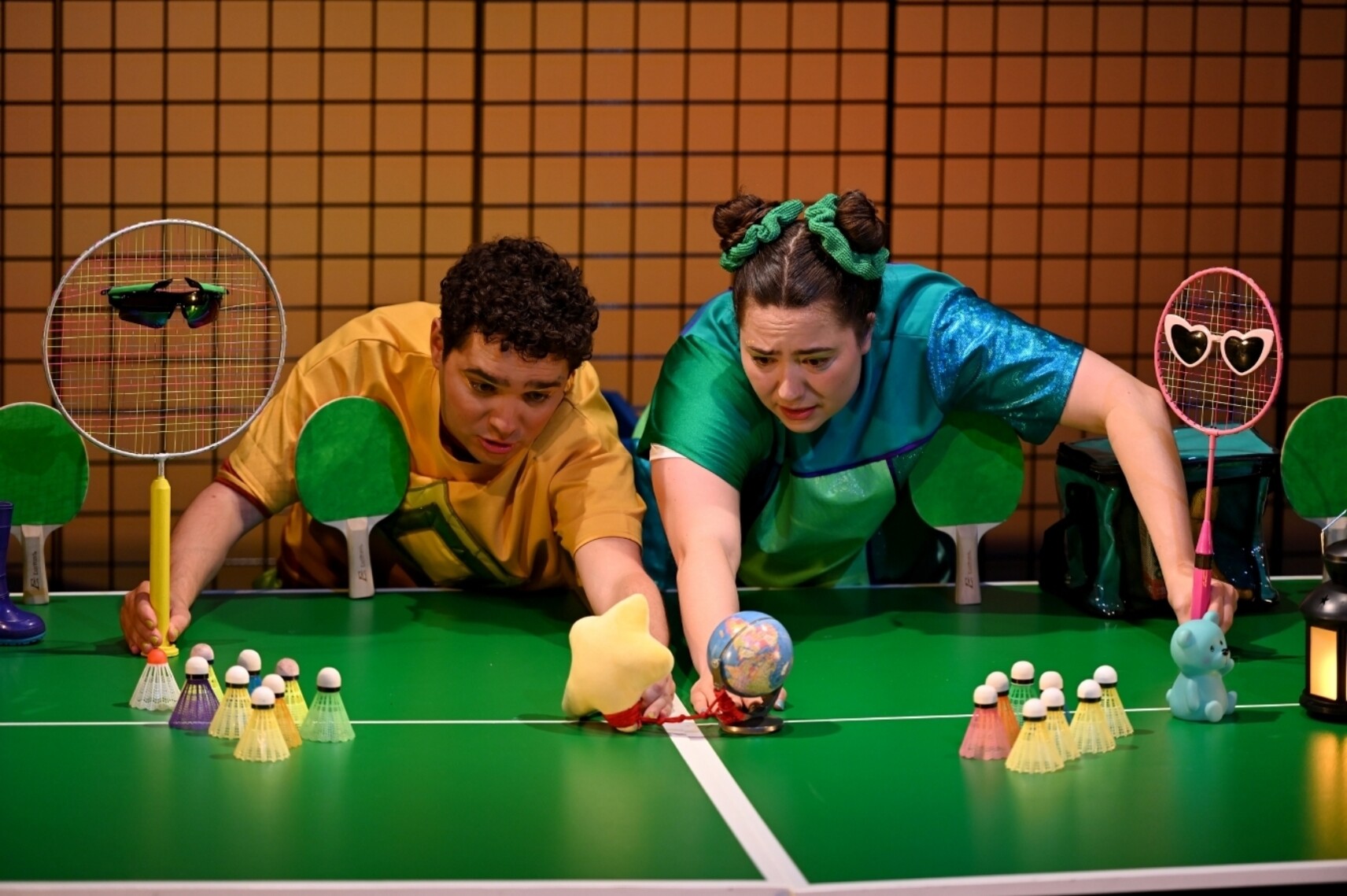 Deux comédien·nes, penché·es sur une table de ping-pong, manipulent des objets miniatures (globe, étoile, figurines et volants de badminton) dans une mise en scène ludique.