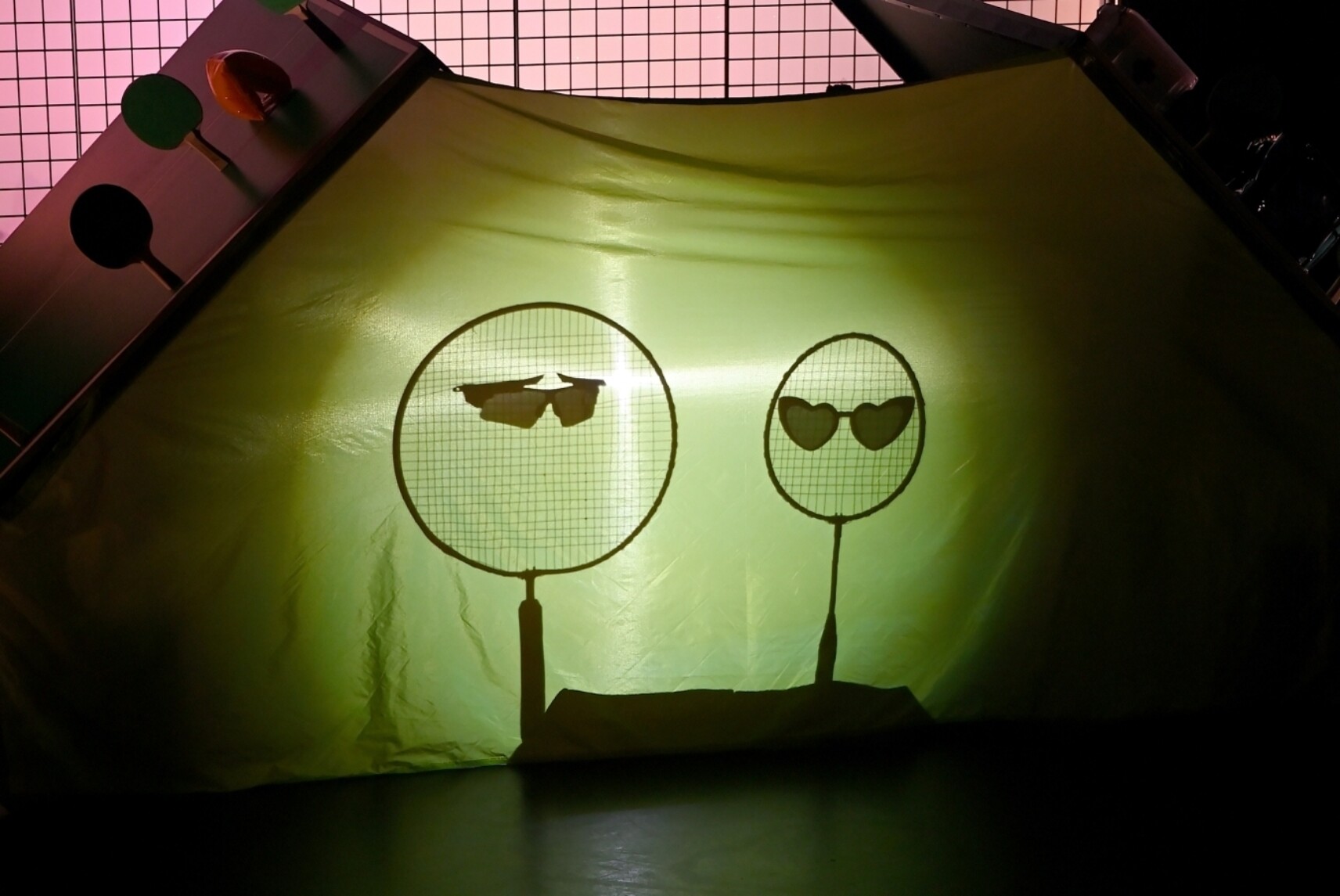 Deux raquettes de badminton projetées en ombre sur un drap vert, l’une portant des lunettes noires et l’autre des lunettes en forme de cœur.