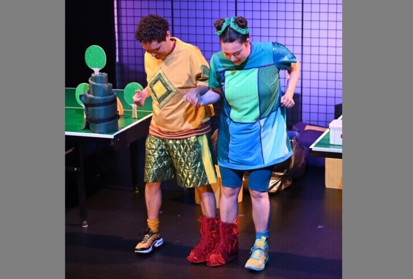 Deux comédien·nes en costumes colorés marchent main dans la main avec un pied fusionné.