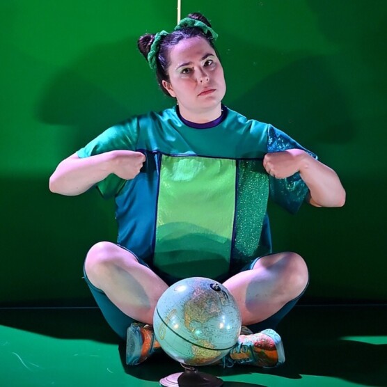Le personnage de Capitaine, en costume bleu et vert, assise en tailleur sur scène avec un globe terrestre devant elle.