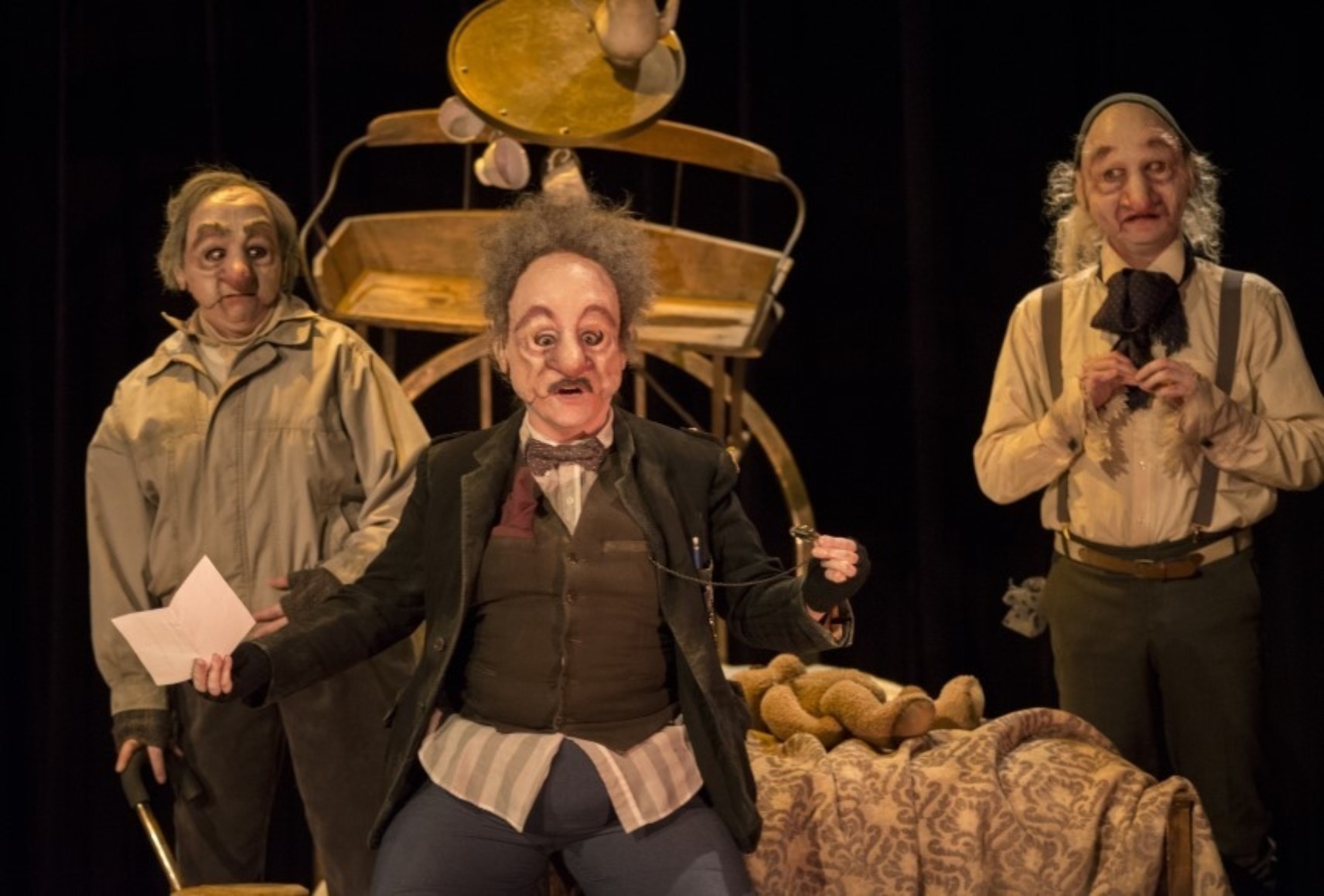 Trois personnages masqués devant une structure de décor. Ils regardent devant eux, vers le public. Le personnage du milieu tient un livre d'une main et de l'autre une montre à gousset. Il semble être en train de parler.