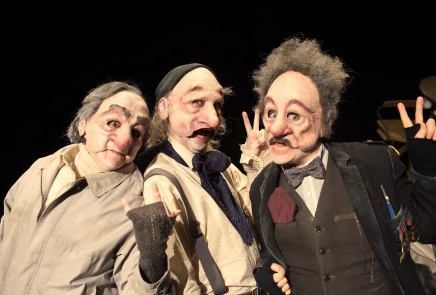 Trois personnages portant des masques d'hommes âgés regardent la caméra en faisant des signes avec leurs doigts, comme des signes de paix. Ils ont des postures dynamiques et sont habillés avec des vêtements beiges, bruns, marine et gris.