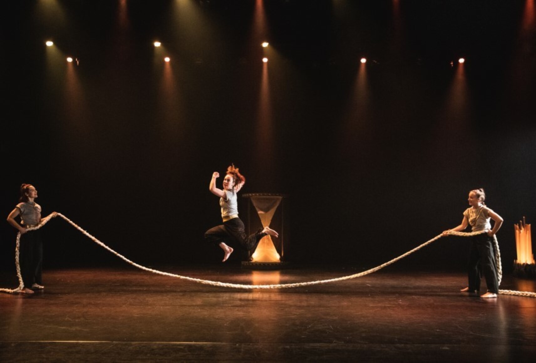 Trois danseuses jouent à sauter à la corde, sur une scène dégagée. En arrière plan, un grand sablier. Elles portent des pantalons noirs et des hauts pâles.