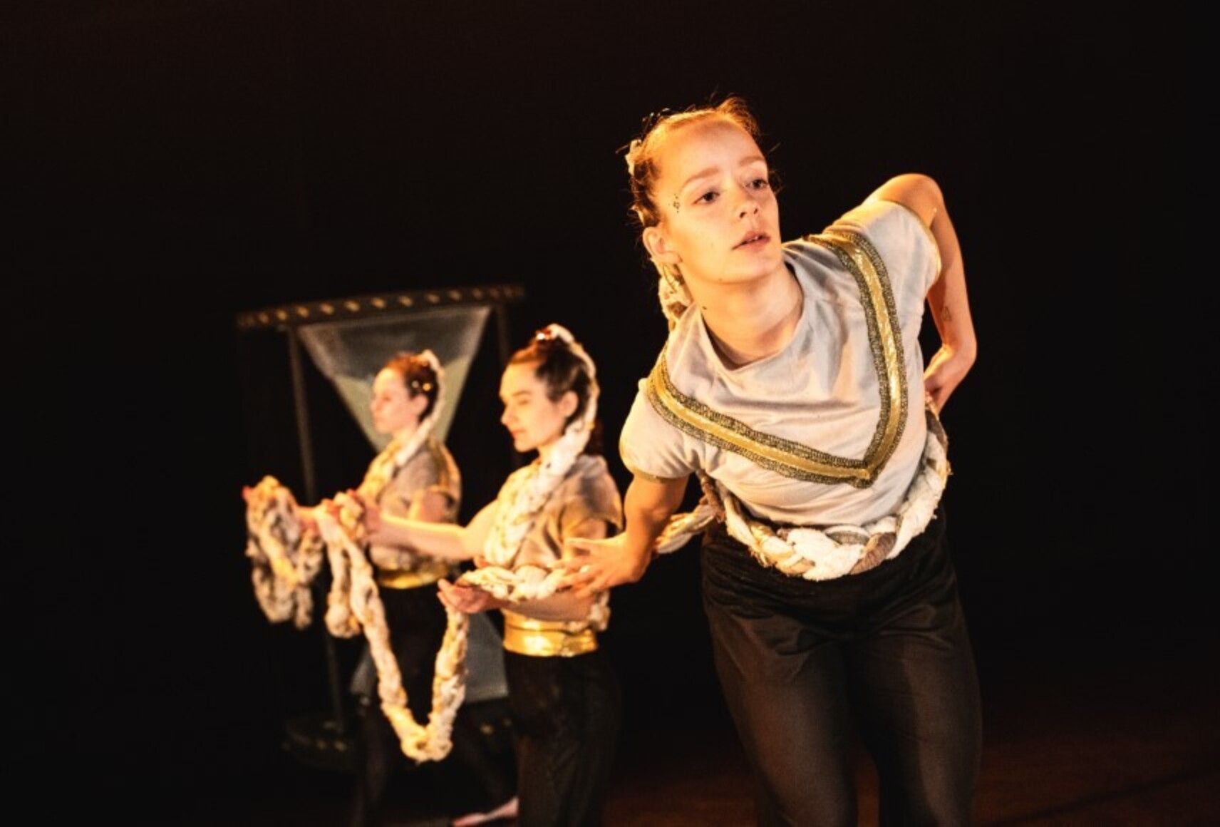 Une danseuse au premier plan avec une très longue tresse s'enroulant autour de sa taille et portée par les deux autres danseuses derrière elle. Elle porte des pantalons noirs et un haut bleu et doré. Au fond, un sablier géant.