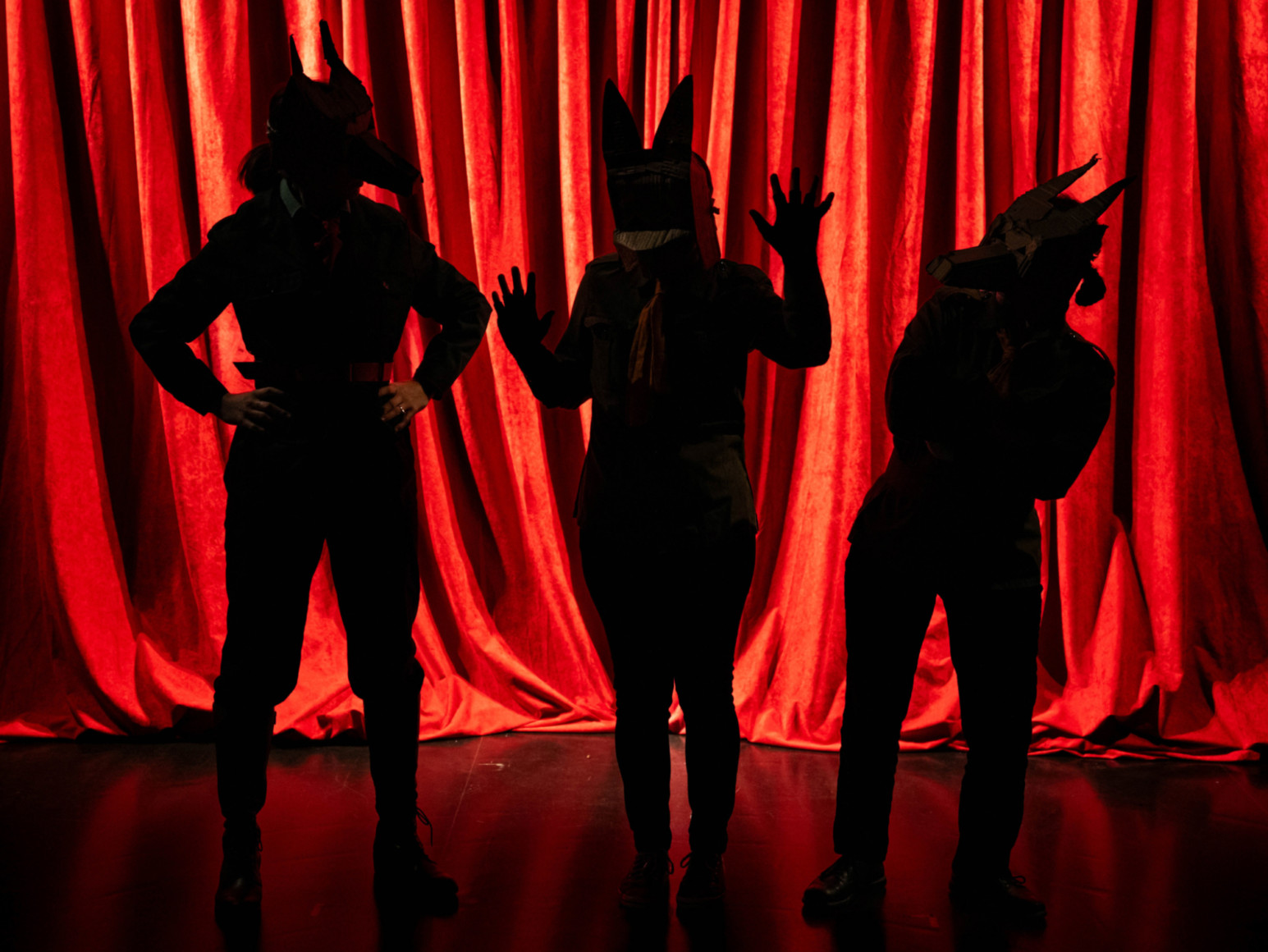 Devant un rideau rouge, photo en contre-jour de trois personnages dont on distingue les masques évoquant des animaux.