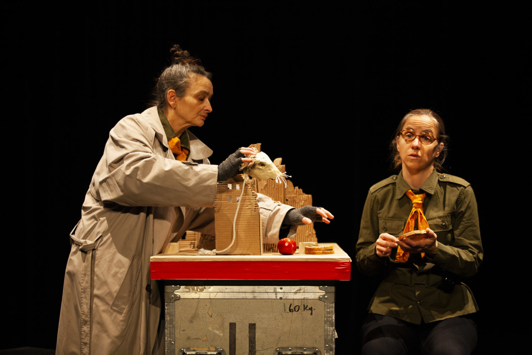 Photo de deux personnages sur fond noir. Celui de gauche porte un long manteau beige et manipule un petit masque de souris. À droite, le personnage avec un habit de scout est assis avec une tranche de pain dans les main.