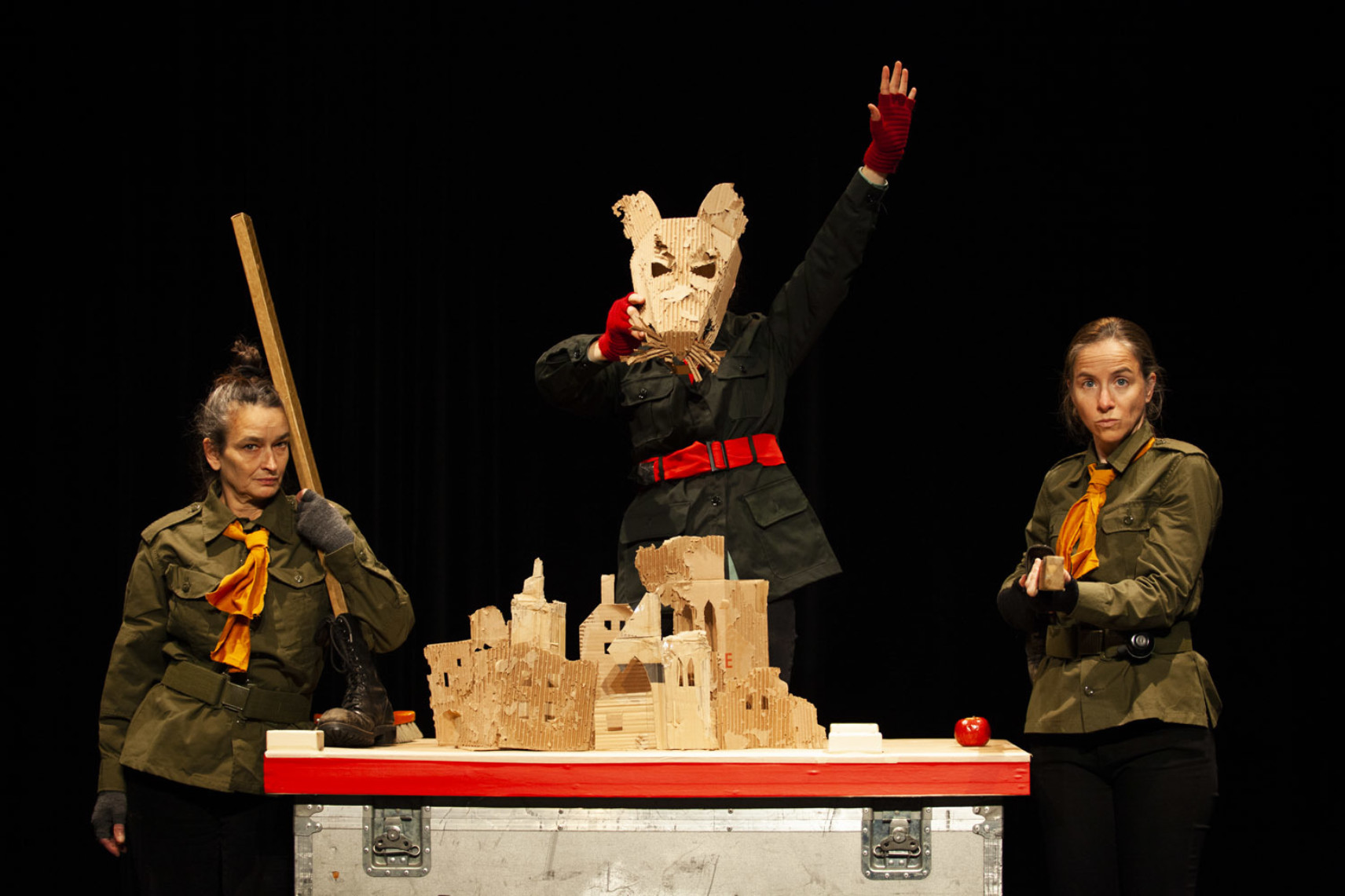 Trois personnage sur scène, habillés avec des vestes de soldat et de scout, devant une maquette de village en carton. Le personnage du milieu porte un masque de souris en carton et lève le bras gauche dans les airs.