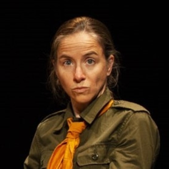 Photo devant fond noir du personnage Félix, portant une chemise kaki et un petit foulard jaune noué au cou. Ses cheveux sont attachés et il regarde doit dans la caméra, les lèvres pincées.