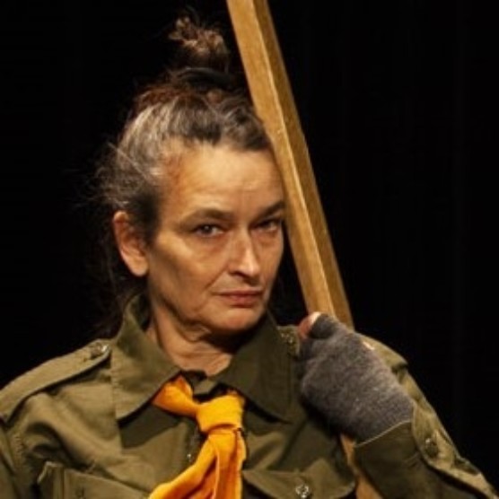 Photo d'un personnage portant une chemise kaki, un petit foulard jaune noué au cou et des gants sans doigts gris. Ses cheveux sont coiffés en chignon et son visage est appuyé sur un bâton de bois qu'il tient dans sa main gauche.