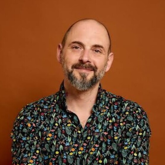Portrait en couleur d'un homme chauve avec une barbe grisonnante, sur fond brun. Il regarde la caméra avec un sourire. Il porte une chemise fleurie.