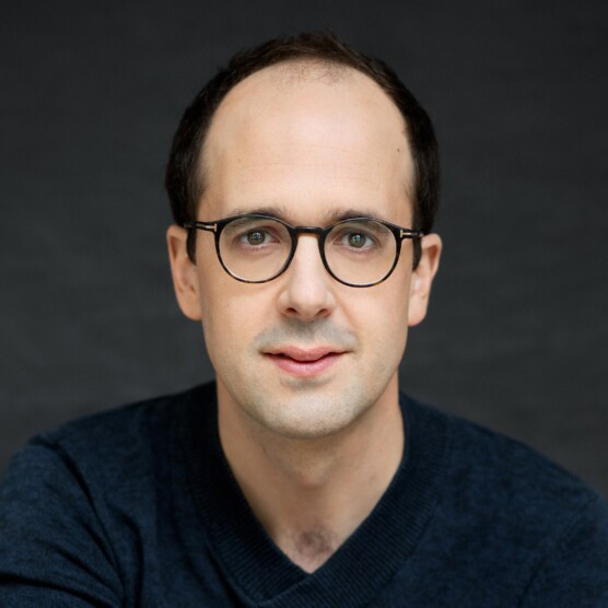 Portrait en couleur d'un homme chauve au teint clair portant des lunettes rondes. Il regarde la caméra. Il porte un chandail bleu foncé.