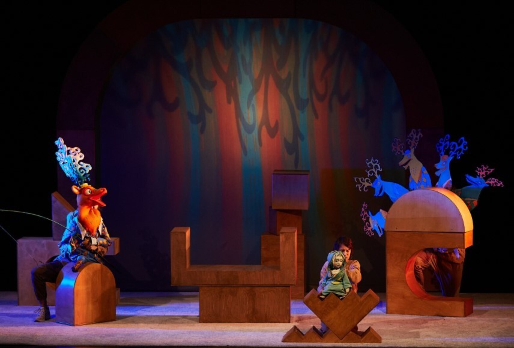 Deux marionnettes, un cerf et un enfant, sont éclairées par des lumières vives, devant un fond coloré. Les éléments de décor sont formés de grandes formes géométriques en bois.