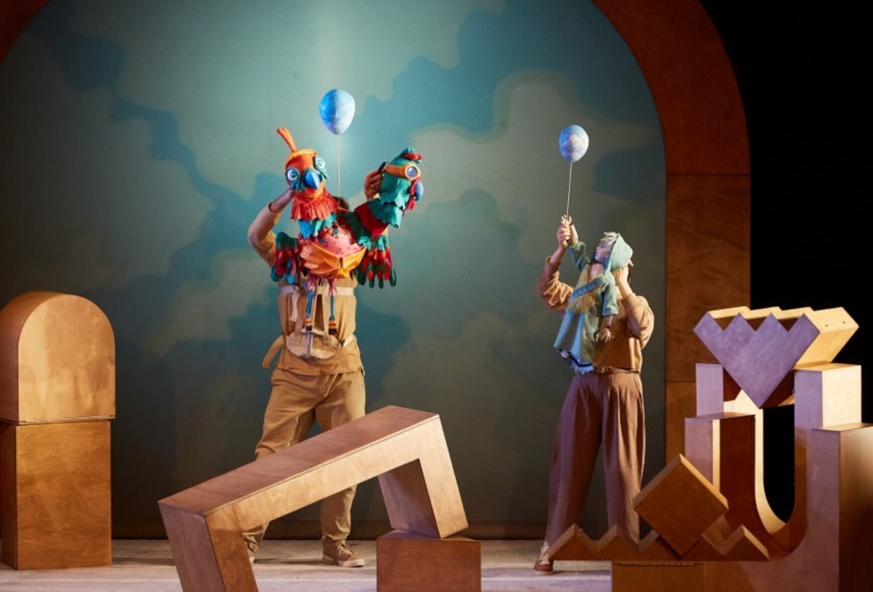 Deux personnes animent des marionnettes, un oiseau à deux têtes et un petit enfant tenant un ballon gonflé bleu, dans un décor de blocs de jeux en bois.