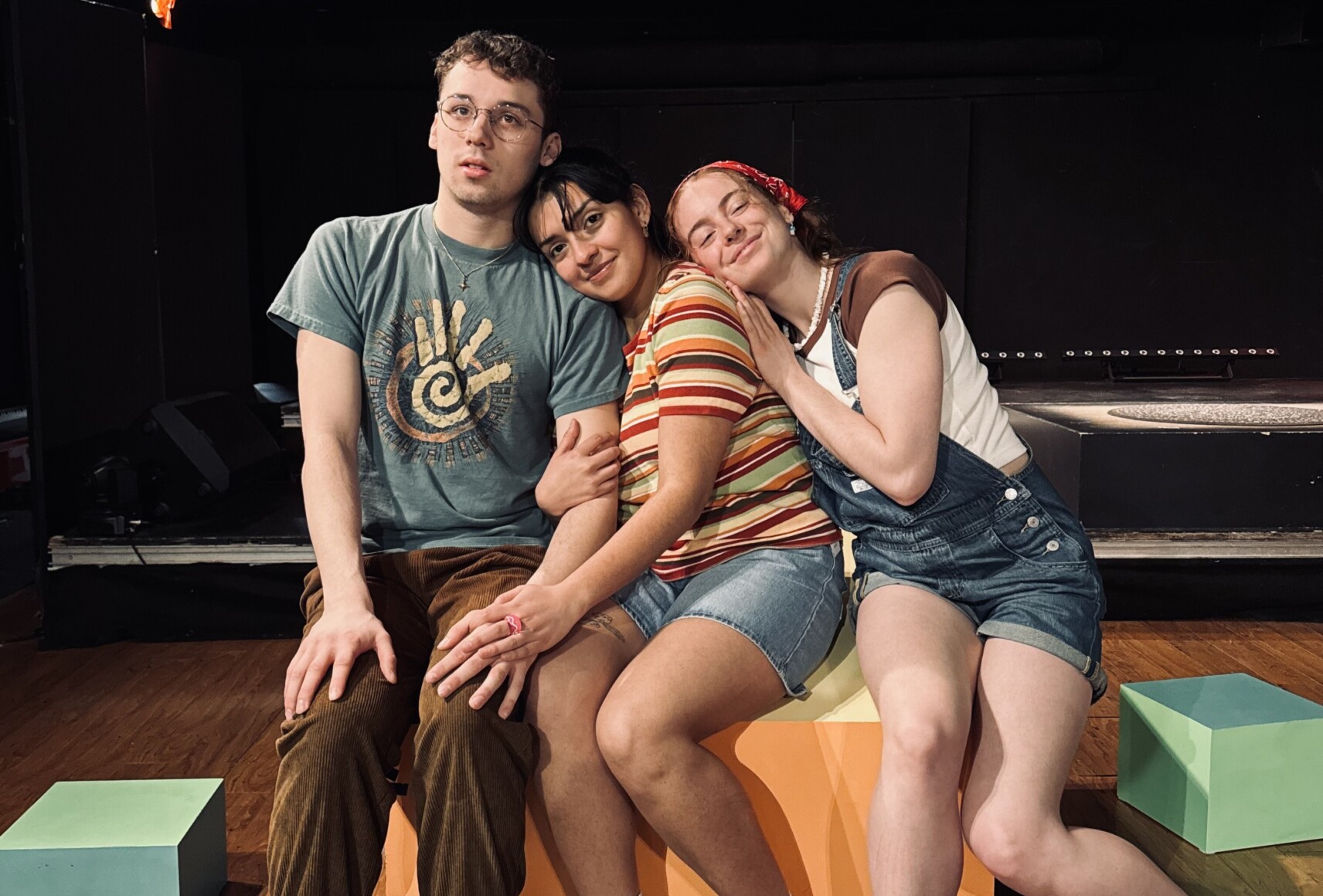 Les trois personnages sont assises côte à côte sur scène, posant sur des cubes colorés.