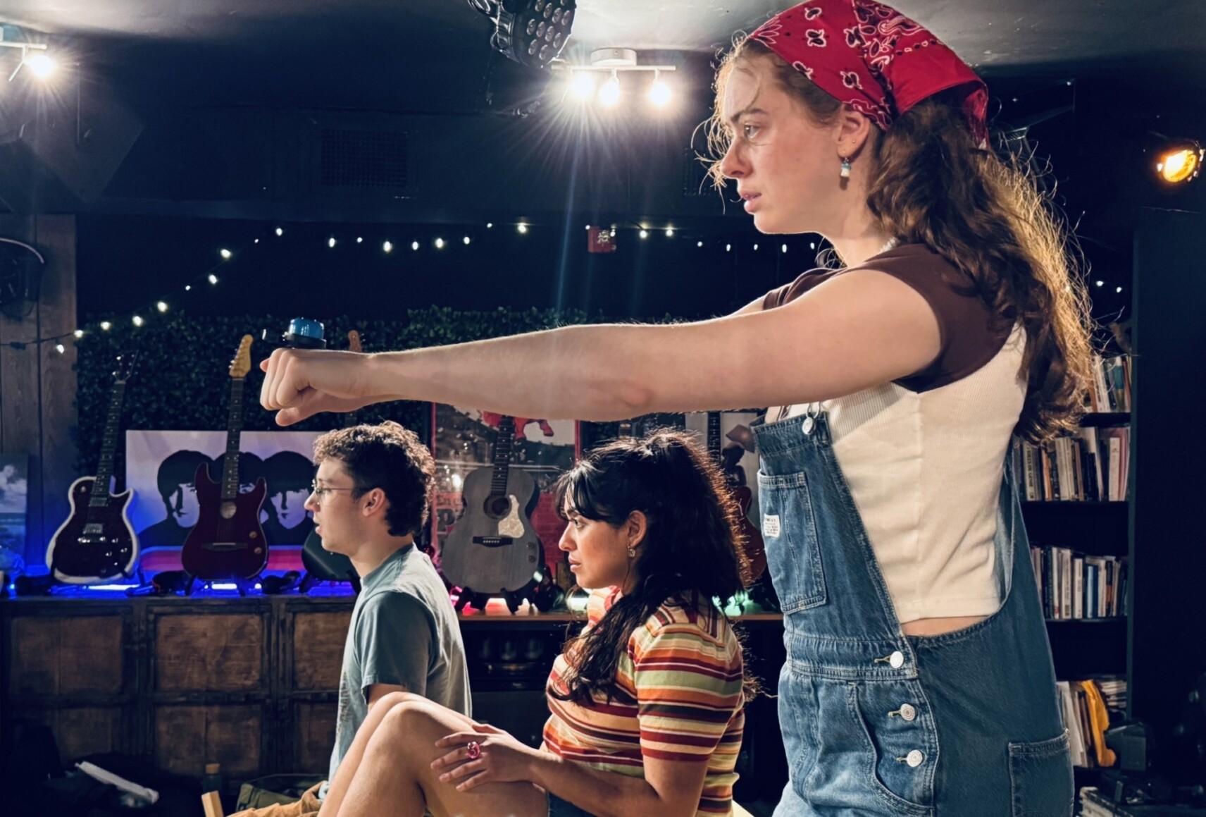 Les trois personnages sont installées sur une plateforme dans une salle décorée de guitares, dont l’une adopte une posture dynamique avec un bras tendu.