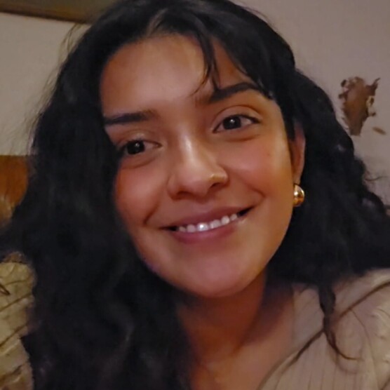 Personne au regard calme et expressif, aux cheveux foncés et bouclés, portant des boucles d’oreilles dorées et un haut beige. Elle est assise sur un canapé en cuir brun, dans un salon chaleureux décoré de livres, de CD et de fleurs séchées.