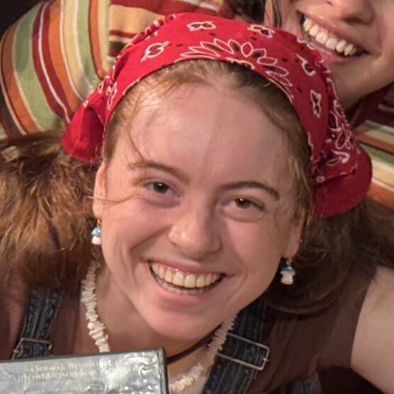 Le personnage de Delphine portant un bandana rouge décoré de motifs blancs est assise devant un coussin rayé aux tons orange, vert et beige. Autour du cou, un collier composé de perles blanches est visible. La personne porte une salopette en denim et un h