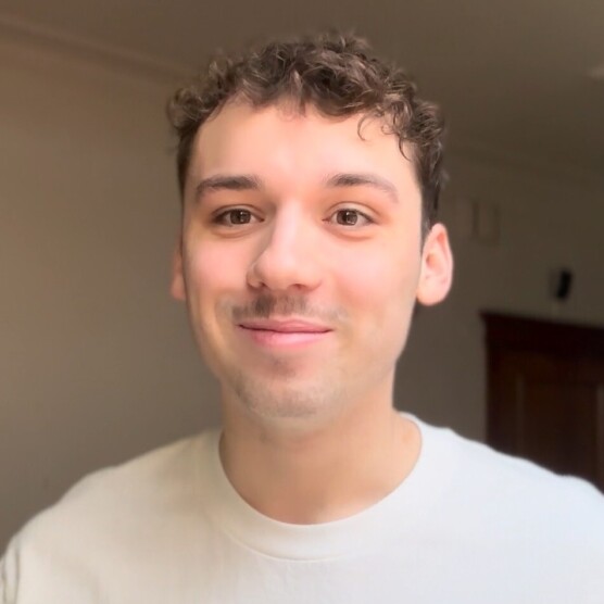 Personne au regard attentif, aux cheveux courts et bouclés, portant un chandail clair. Elle se tient dans une pièce lumineuse à l’arrière-plan neutre, où l’on aperçoit une porte en bois et un coin télévision.