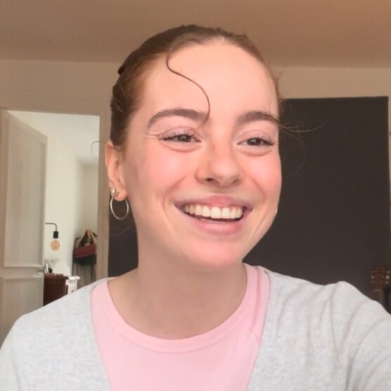 Personne souriante, les cheveux attachés avec une mèche bouclée sur le front, portant un chandail rose sous un cardigan gris pâle. L’arrière-plan montre une pièce bien éclairée avec des étagères murales.