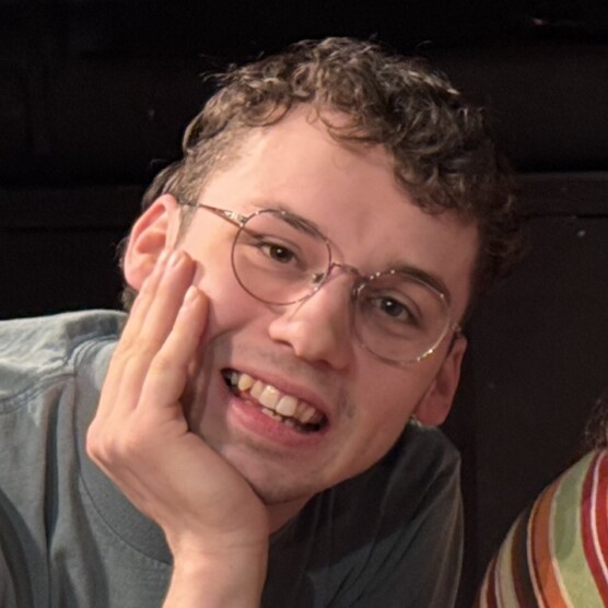 Le personnage de Mowgli est assis dans un environnement intérieur sombre, appuyée sur une main placée sous la mâchoire. La personne porte un t‑shirt uni et se trouve légèrement inclinée vers l’avant. À droite, on distingue une partie d’un coussin ou d’un