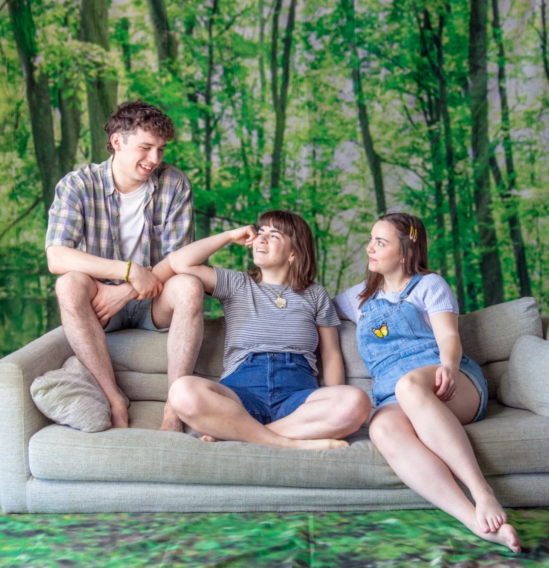 Trois jeunes sont assis·es sur un canapé beige, devant un décor de forêt. Elles et ils échangent des regards et sourient, dans une posture détendue et conviviale, évoquant une discussion complice.