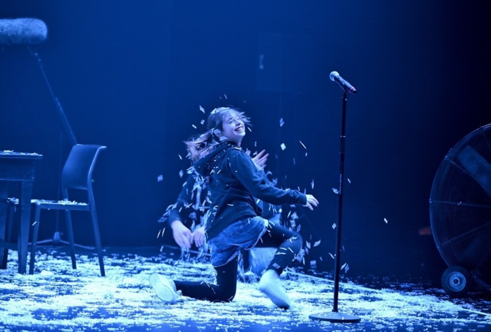 Dans une lumière bleutée, Éléonore rit dans la fausse neige, agenouillée au sol. Elle porte des collants noirs, des shorts en jeans et un coton ouaté noir. Un micro sur pied, une chaise et une table sont visibles sur scène.