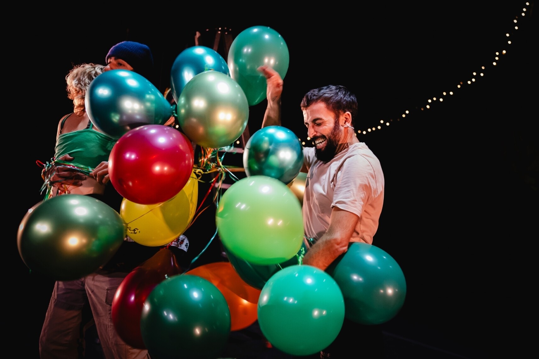 Photo d'un gigantesque bouquet de ballons colorés et visiblement emmêlés qui prend toute la place. Un personnage masculin semble se débattre et essayer de le contenir avec un très grand sourire aux lèvres.