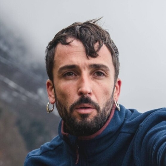 Photo d'un jeune homme, les cheveux courts et portant la barbe, en plein mouvement dans un décor de montagnes. Il a des piercings au nez et aux oreilles.