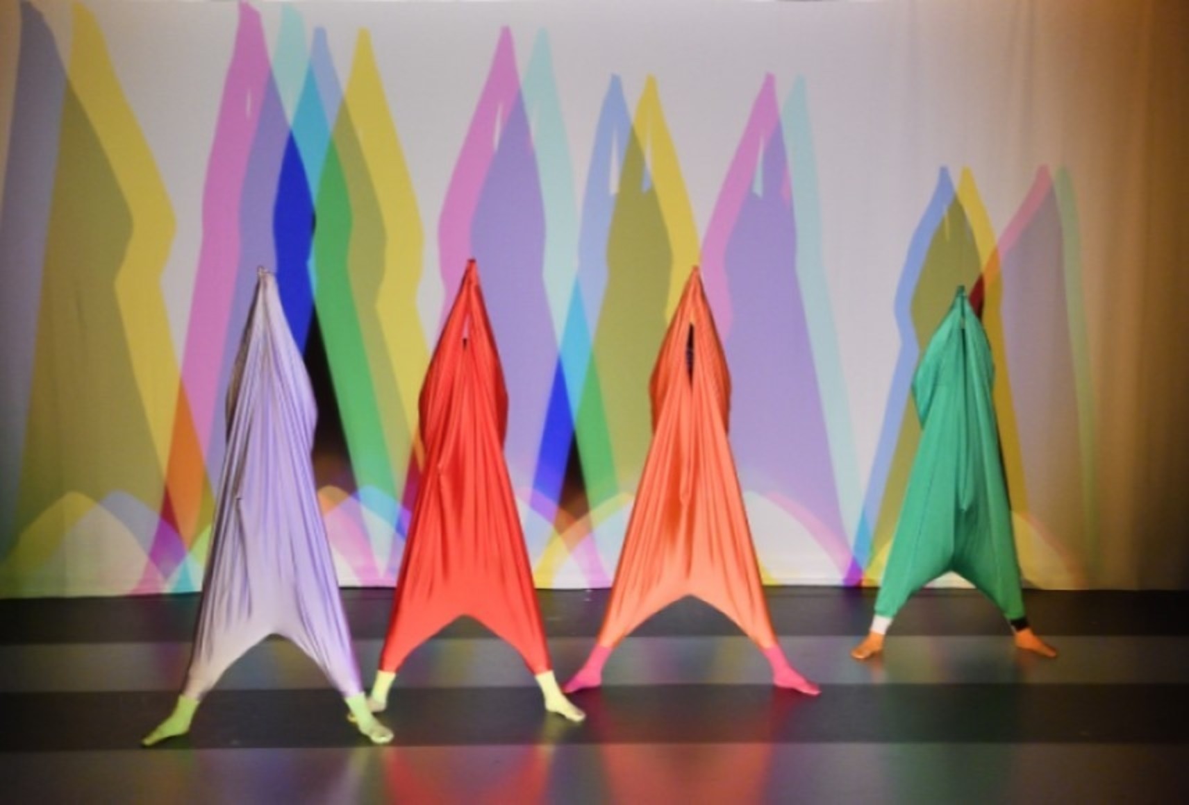 Quatre danseuses en costumes colorés projettent des ombres de couleur triangulaires derrière elles.