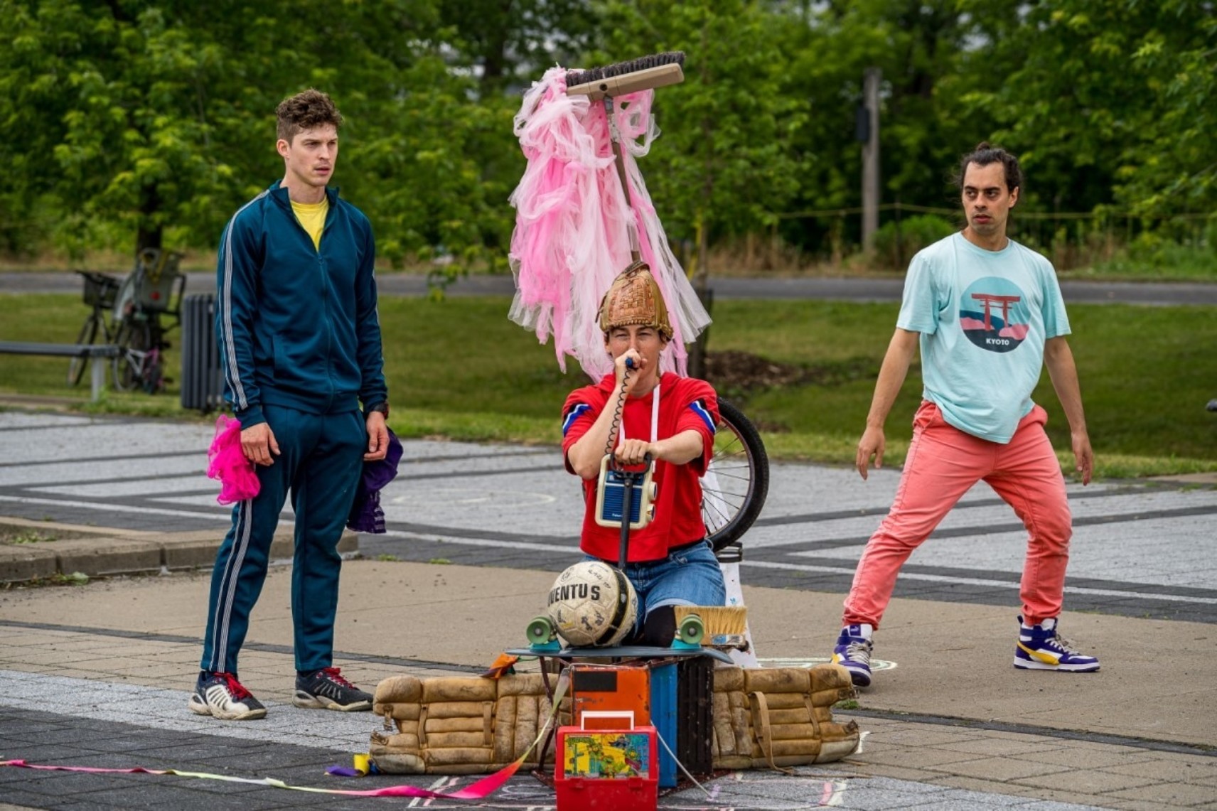 Photo en extérieur de trois personnages en habits colorés. Le personnage au centre en avant-plan est assis dans une structure faite d'équipement sportif et d'un balais dressé avec une jupe en tulle. Les deux autres personnages en arrière-plan le regardent